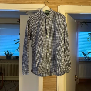 Blårutig skjorta Tommy Hilfiger L - Klassisk blårutig skjorta från Tommy Hilfiger i storlek L. Skjortan har lång ärm, knappar framtill och button down-krage. Tillverkad i mjuk bomull som känns skön mot huden. Perfekt för dig som gillar en stilren och tidlös look.