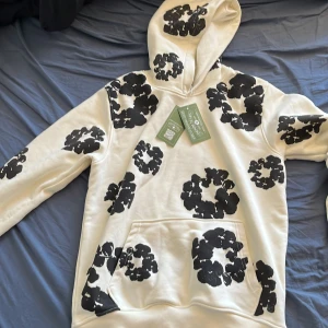 Vit hoodie med svarta blommor Denim Tears - Säljer en vit hoodie från Denim Tears i storlek S med stora svarta blommönster över hela tröjan. Hoodien har huva, känguruficka och är tillverkad i mjuk bomull. Perfekt statement-plagg för dig som gillar unik streetwear. Priset kan diskuteras😁