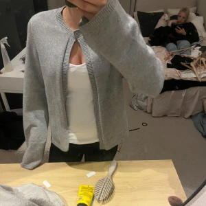 Ljusgrå kofta med knapp - Superenkel och stilren ljusgrå kofta med rund hals och en knapp upptill. Koftan är långärmad och har ribbade muddar och kant. Perfekt att slänga över en t-shirt för en chill look.