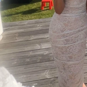 Vit spetsklänning med blommönster - Supersnygg långklänning i vit spets med blommigt mönster och figurnära passform. Klänningen har en lätt champagnefärgad underton, är axelbandslös och har fina rynkade detaljer längs hela längden. Perfekt för dig som vill sticka ut med en elegant look.