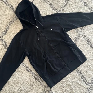 Svart hoodie från Polo Ralph Lauren - Svart hoodie från Polo Ralph Lauren med klassisk vit broderad logga på bröstet. Tröjan har dragkedja framtill, huva med snörning och två fickor. Tillverkad i mjuk bomull, perfekt för en chill och stilren look. För frågor som måttet av tröjan skicka ett PM.