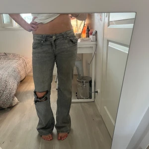 Gråa bootcut jeans med slitning - Säljer ett par gråa jeans med bootcut passform och snygg slitning på ena knät. Jeansen har låg midja, raka ben och coola bakfickor med knappdetalj. Perfekta för en avslappnad och trendig look.