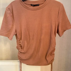 Beige ribbad topp med snörning Zara - Säljer en beige ribbad topp från Zara i storlek M. Toppen har korta ärmar, rund hals och snygg snörningsdetalj i sidan som ger en cool, lite croppad look. Perfekt för dig som gillar bekväma plagg med det lilla extra.