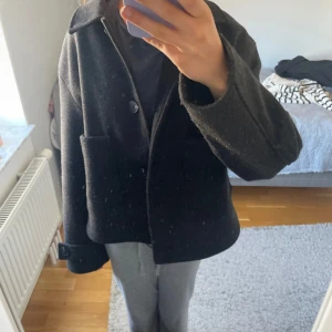 Svart ulljacka med stora fickor - Säljer en svart, boxig ulljacka med klassisk krage och stora utanpåfickor. Jackan har knäppning framtill med stora knappar och långa ärmar med snygg detalj vid ärmslutet. Perfekt för dig som gillar en stilren och enkel look. Lite nopprig 