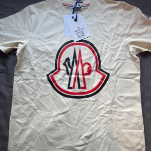 Beige Moncler t-shirt med stor logga - Snygg beige t-shirt från Moncler med stor, ikonisk logga i svart och rött på bröstet. Klassisk passform och rund halsringning. Tillverkad i mjuk bomull som känns skön mot huden. Perfekt för dig som gillar stilrena och exklusiva plagg med statement-tryck.