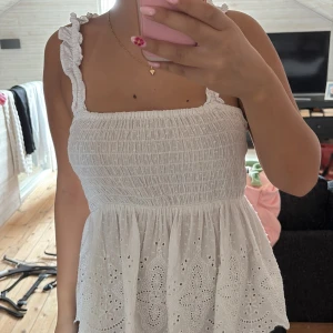 Vit broderad blus med volangband - Supersöt vit blus med smockad överdel och breda axelband med volangdetaljer. Nederdelen är utsvängd med broderade blommor och hålmönster, perfekt för sommarens alla dagar. Lätt och luftig känsla!