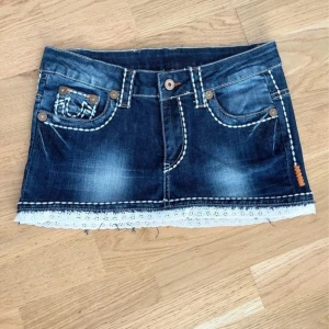 True Religion jeanskjol med spetskant - Supersnygg kort jeanskjol från True Religion med coola kontrastsömmar och detaljerade bakfickor. Kjolen har en vit spetskant längst ner som ger en extra touch. Klassisk blå denim med slitningar och fickor fram och bak. Perfekt för dig som vill sticka ut!