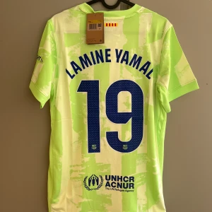 FC Barcelona Lamine Yamal 19 Nike tröja - Säljer en FC Barcelona fotbollströja med Lamine Yamal och nummer 19 på ryggen. Tröjan är ljusgrön med vita detaljer, har korta ärmar och är tillverkad i ett lätt, ventilerande material. Nike-logga, Spotify och UNHCR-tryck samt klubbmärke på bröstet.