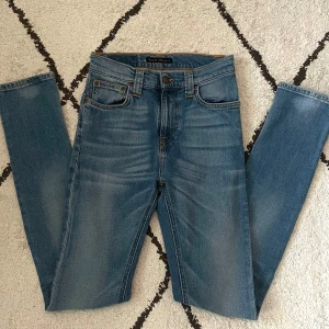 Blå Nudie Jeans, rak modell W27 L32 - Säljer ett par klassiska blå jeans från Nudie Jeans med snygga kontrastsömmar och broderade bakfickor. Modellen har normal midja och raka ben, tillverkade i mjuk bomullsdenim. Perfekta för dig som gillar en tidlös och clean look.