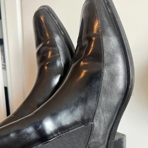 Svarta chelsea boots i skinn - Snygga svarta chelsea boots med klassisk rund tå och elastiska paneler på sidorna. Skorna är tillverkade i blankt skinn och har en låg klack. Perfekta för dig som gillar stilrena och tidlösa skor.