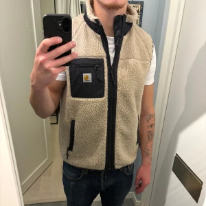 Beige fleeceväst från Carhartt WIP - Säljer en beige fleeceväst från Carhartt WIP med svarta detaljer. Västen har hög krage, dragkedja framtill och tre fickor med svarta kanter.