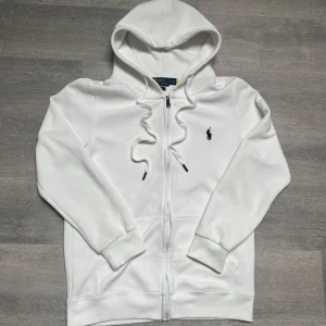 Vit hoodie från Polo Ralph Lauren - Snygg vit hoodie från Polo Ralph Lauren med broderad logga på bröstet. Tröjan har huva med snörning, dragkedja framtill och två fickor. Tillverkad i mjuk bomull och har en clean, klassisk look som passar perfekt till jeans eller joggers.