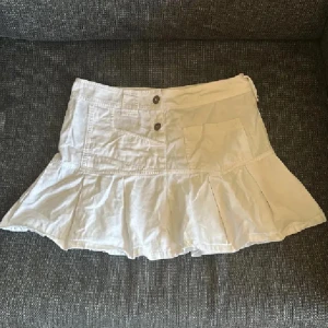 Vit volangkjol med fickor - Supersöt vit kortkjol med volangkant och två stora knappar framtill. Kjolen har praktiska fickor och är sydd i ett lätt bomullstyg, perfekt för varma dagar. Sitter snyggt i midjan och ger en fräsch och trendig look. Tryck ej köp nu