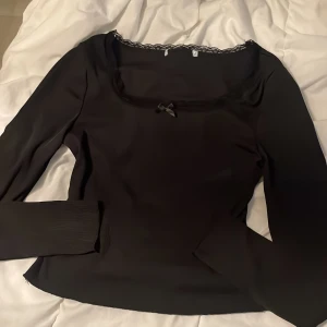 Svart ribbad långärmad topp med spets - Svart långärmad topp med fyrkantig ringning, dekorerad med spetskant och en liten rosett framtill. Materialet är ribbat och stretchigt vilket ger en snygg passform. Perfekt för dig som gillar stilrena detaljer och en feminin touch.