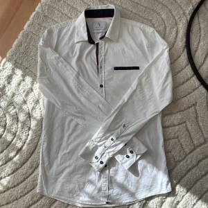 Vit skjorta från Savvy Citizen, L - Vit skjorta från Savvy Citizen i storlek L. Skjortan har svarta detaljer på krage, knappslå och bröstficka samt svarta knappar. Tillverkad i bomull och har lång ärm med dubbla knappar vid manschetten. Snygg kontrast med röd linje på insidan av kragen.