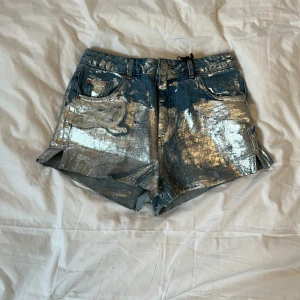 Silverglittriga jeansshorts Topshop - Unika jeansshorts från Topshop med hög midja och glittrig silverfolie över hela tyget. Klassisk femficksmodell med råa benslut och cool metallic look. Perfekta för dig som vill sticka ut och älskar statement-plagg.