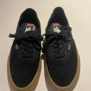 Svarta Vans sneakers med gummisula - Svarta Vans popcush sneakers i klassisk modell med lågt skaft, snörning och brun gummisula. Ovandelen är i mocka och textil, med diskreta rutiga detaljer vid snörningen. Perfekta för skate eller streetstyle. Näst intill oanvända.