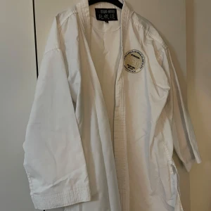 Vit karatejacka från Budo-Nord 180 - Karatejacka från Budo-Nord i vitt, klassisk modell med broderad patch på bröstet. Tillverkad i slitstark bomull, perfekt för träning eller tävling. Jackan har långa ärmar och traditionell krage. Snygg och enkel design för dig som kör kampsport. (Byxor och jackan)