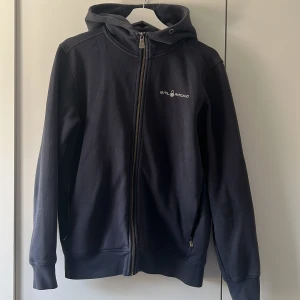 Svart hoodiejacka från Sail Racing - Svart hoodiejacka från Sail Racing med dragkedja hela vägen, huva och diskret logga på bröstet. Hoodien har långa ärmar och två fickor med dragkedja. Perfekt för dig som gillar en clean och sportig stil.