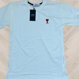 Vit t-shirt från Ami Paris  - Vit t-shirt från Ami Alexandre Mattiussi i 100% . Klassisk passform med korta ärmar och rund hals. På bröstet finns ett broderat rött hjärta med bokstaven A. Perfekt basic med snygg detalj som sticker ut.