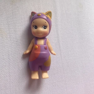 Sonny Angel - Bilderna visar en söt leksaksfigur med lila overall och kattöron, men ingen bok syns på bilderna. Ingen information om titel, författare eller innehåll kan utläsas.