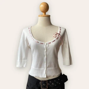 Vit kofta med rosa rosett från OFUON palette - Supersöt vit kofta från OFUON palette med trekvartsärm och knappar framtill. Urringningen är dekorerad med ett rosa band och en liten rosett som ger en gullig vibe. Modellen är croppad och sitter tajt mot kroppen. Perfekt för dig som gillar en feminin och söt stil. Importerad från Japan. Jfashion Lolita kawaii coquette kofta. 