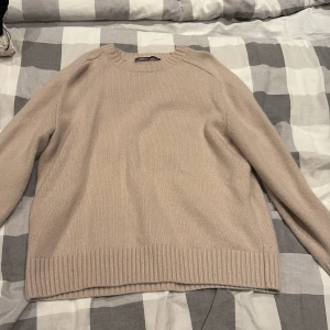 Beige stickad tröja från Zara - En stilren beige stickad tröja från Zara i storlek M. Tröjan har rund halsringning, långa ärmar och breda ribbade muddar vid ärmslut och nederkant. Perfekt för lager-på-lager och enkel att matcha med jeans eller kjol.