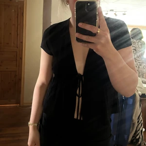 Svart knyt från Gina Tricot - Svart omlottopp från Gina Tricot med djup v-ringning och knyt framtill. Toppen har korta ärmar och en lätt rynkad midja som ger en snygg siluett. Perfekt för dig som gillar en trendig och stilren look.