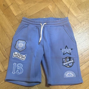 Blå college-shorts från BoohooMAN - Blå mjuka shorts från BoohooMAN med coola broderade patches och tryck, bland annat siffror, stjärna och text som 'Worldwide' och 'Limited Edition'. Shortsen har snörning i midjan och två fickor. Perfekta för en avslappnad och sportig stil.