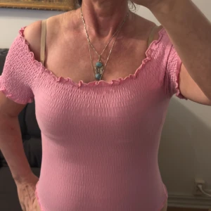 Rosa offshoulder topp med volangkant - Supersöt rosa offshoulder topp med smockad struktur och volangkant längs halsringning, ärmslut och nederkant. Toppen är kortärmad och har en tight passform som framhäver axlarna. Tillverkad i mjuk bomull med lite stretch för extra komfort.