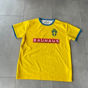 Sverige fotbollströja gul Bauhaus nr 19 - Gul fotbollströja från Sveriges landslag med blå detaljer vid krage och ärmslut. Bauhaus-logga på bröstet och ryggen, samt stort nummer 19 på ryggen. Broderat landslagsemblem på bröstet. Tillverkad i ventilerande polyester. Storlek S/M. Perfekt inför EM just nu.