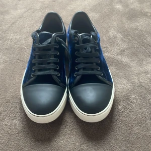 Blå sneakers från Lanvin - Snygga sneakers från Lanvin i mörkblå med svarta snören och tåparti i matt svart. Skorna har vit sula och klassisk rund tå. Perfekta för dig som vill sticka ut med exklusiva material och cool design.