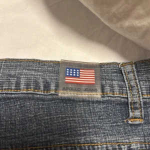 Jeanskjol från Apollo Jeans - Snygg jeanskjol från Apollo Jeans i klassisk blå denim med coola kontrastsömmar i gult. Kjolen har fem fickor, bälteshällor och dragkedja framtill. Bakfickorna har dekorativa broderier som ger extra edge till looken.