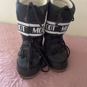 Svarta Moon Boot snowboots - Svarta Moon Boot snowboots med vit logga och breda snören framtill. Skorna har en hög, vadderad design och vadderad inifrån. Även fast sulan är försliten så syns det inte bakifrån eller när man går, men jag har ändå tagit hänsyn till det när jag lagt priset.