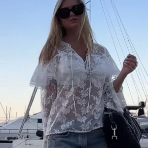Vit blommig spetsblus med volang - Superfin transparent vit blus i spets med blommigt mönster och volangdetaljer över axlarna. Blusen har långa ärmar, knytband vid halsen och romantisk vibe. Perfekt att styla med jeans eller kjol för en trendig look.                        LITEN I STORLEKEN