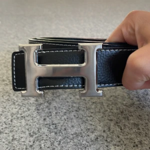 Svart H3rmès skärp med H-spänne - Säljer ett svart skärp från H3rmès med ikoniskt H-spänne i silverfärgad metall. Bältet är gjort i  skinn med vita kontrastsömmar längs kanterna. Perfekt accessoar för att lyfta din outfit med en lyxig touch.