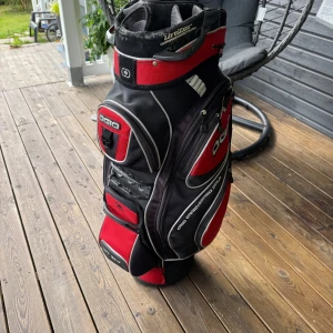 OGIO svart och röd golfbag - Säljer en sportig golfbag från OGIO i svart och rött med flera smarta fack och dragkedjor. Väskan har robust bärrem, tydliga OGIO-loggor och gott om plats för klubbor och tillbehör. Perfekt för dig som vill ha stil och funktion på golfbanan.