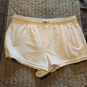 Vita Juicy Couture shorts med strass - Superfräscha vita shorts från Juicy Couture i mjuk velour med elastisk midja och snörning. Snygga dragkedjor på fickorna och ikonisk Juicy-text i strass på baksidan. Perfekta för sommaren och chill dagar.