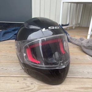 Moped hjälm  - Snygg svart MC-hjälm från LS2 med blank finish och röd mesh på insidan. Hjälmen har ett stort visir och smart spänne under hakan. Perfekt för dig som vill ha både stil och säkerhet på vägen. Mopedhjälm i storlek M. Tvättas innan jag postar 