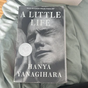 A Little Life - Säljer romanen 'A Little Life' av Hanya Yanagihara. Omslaget är svartvitt med ett porträtt av en man och texten är i stora vita bokstäver. Boken är en Man Booker Prize-finalist och har en sticker på framsidan.