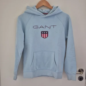Ljusblå hoodie från Gant - Ljusblå hoodie från GANT med broderad logga och sköld på bröstet. Tröjan har huva, magficka och långa ärmar. Perfekt för en avslappnad stil och enkel att matcha med jeans eller mjukisbyxor. Perfekt till sommaren. Strl 150-160cm.
