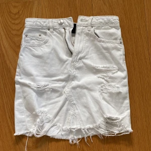 Vit jeanskjol med slitningar från H&M Divided - Säljer en vit kort jeanskjol från H&M Divided i storlek XS. Kjolen har slitna detaljer, fransig nederkant och klassiska fem fickor. Perfekt för dig som gillar en edgy och avslappnad stil.