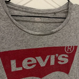 Grå t-shirt från Levi's med logga - Säljer en klassisk grå t-shirt från Levi's med stor röd logga framtill. T-shirten har rund halsringning och korta ärmar. Perfekt för en avslappnad och stilren look.