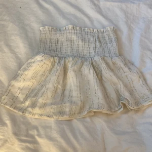 Vit kortkjol med glittriga ränder - Supersöt vit kortkjol med bred smock i midjan och glittriga ränder. Kjolen har en luftig och lätt känsla, perfekt för varma dagar. Den stretchiga midjan gör den bekväm och enkel att bära.