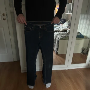 Mörkblå jeans från Nudie Jeans - Säljer ett par klassiska mörkblå jeans från Nudie Jeans med orangea kontrastsömmar och femficksdesign. Byxorna har raka ben och normal passform, samt läderpatch med logga bak i midjan.