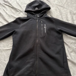 Svart hoodiejacka från Sail Racing - Svart hoodiejacka från Sail Racing med dragkedja framtill och en bröstficka med dragkedja. Jackan har en broderad logga på huvan och tryckt logga på bröstet. Perfekt för en sportig och avslappnad stil.