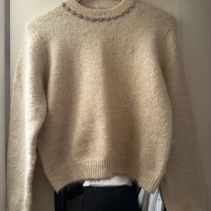Beige stickad tröja med pärldetaljer - Säljer en beige stickad tröja med pärlor runt halsringningen. Tröjan har lång ärm och ribbade muddar vid ärmslut och nederkant. Perfekt för dig som vill ha en enkel men ändå unik look.