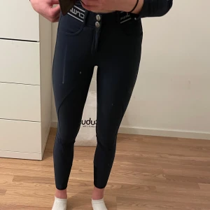 Mörkblå ridbyxor från CRW - Snygga mörkblå ridbyxor från CRW med dubbla knappar i midjan och elastisk logotypresår. Byxorna har en tight passform och är perfekta för ridning. Klassisk design med fickor framtill och förstärkta detaljer.