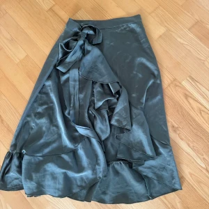 Grön omlottkjol med volang - Säljer en snygg grön omlottkjol i satinliknande material. Kjolen har volangdetaljer längs kanten och knyts i midjan med en rosett. Perfekt för dig som vill ha en trendig och elegant look.