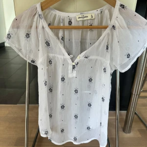 Vit blus med blommigt mönster från Abercrombie - Söt och luftig vit blus från Abercrombie med små svarta blommor. Blusen har korta ärmar, v-ringning med knappar och ett lätt transparent tyg. Är storlek M barn som är en vuxen XS ungefär 